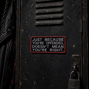 May include: A rectangular patch with the text "JUST BECAUSE YOU'RE OFFENDED, DOESN'T MEAN YOU'RE RIGHT." in white letters on a black background with a red border. The patch is affixed to a dark, textured surface, with a padlock visible.