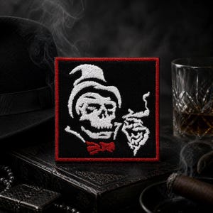 May include: Square black patch with a white skull wearing a hat and bow tie, smoking a cigar. The patch has a red border and is set against a dark background with a hat, glass, and cigar.