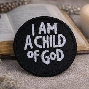 May include: A black circular patch with the white embroidered text "I AM A CHILD OF GOD." The patch is set against a backdrop of an open book, a wooden cross, and a lit candle, suggesting a religious theme.