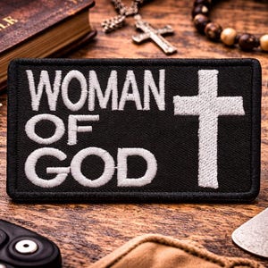 May include: A black patch embroidered with the words "WOMAN OF GOD" and a white cross. The patch is surrounded by religious items, including a Bible, crosses, and jewelry, all arranged on a wooden surface.