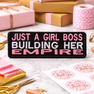 May include: A black embroidered patch with the text "JUST A GIRL BOSS BUILDING HER EMPIRE" in white and pink. The patch is rectangular and sits on a marble surface, surrounded by gift boxes, scissors, and stickers.