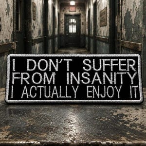May include: Black rectangular patch with white embroidered text: "I DON'T SUFFER FROM INSANITY I ACTUALLY ENJOY IT." The patch is on a distressed metal surface. The background shows a hallway with peeling paint and the words "THE MADNESS".