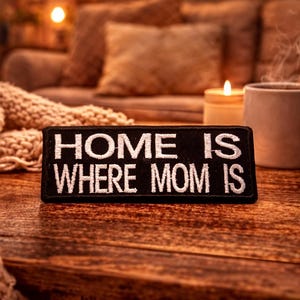 Może przedstawiać: Czarna prostokątna naszywka z białym napisem "HOME IS WHERE MOM IS". Leży na drewnianym stole, obok parującego kubka, zapalonej świecy i oprawionego zdjęcia. Przytulna, ciepła scena.