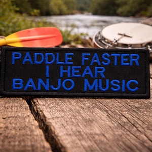 May include: Black patch with blue embroidered text that reads "PADDLE FASTER I HEAR BANJO MUSIC." A red and yellow paddle and a banjo are in the background, resting on a wooden surface.