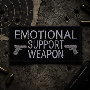Può includere: Toppa rettangolare nera con la scritta bianca "EMOTIONAL SUPPORT WEAPON", con un'immagine di pistola grigia su entrambi i lati. La toppa è su una superficie scura e strutturata con bossoli e un bersaglio sullo sfondo.