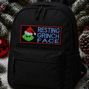 May include: Black backpack with a front pocket and a mesh side pocket. The backpack features an embroidered patch with the Grinch wearing a Santa hat and the words "RESTING GRINCH FACE" in blue and red.