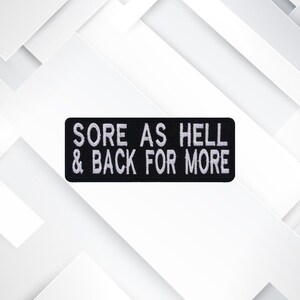 May include: A black rectangular patch with white embroidered text that reads "SORE AS HELL & BACK FOR MORE". The patch is likely designed to be sewn or ironed onto clothing or accessories.