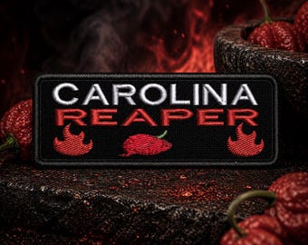 Könnte beinhalten: Ein schwarzer Aufnäher mit den Worten "CAROLINA REAPER" in Weiß und Rot. Der Aufnäher zeigt eine rote Chilischote und Flammen. Im Hintergrund befinden sich mehrere rote Chilischoten.