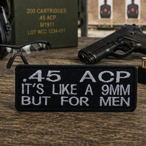 Puede incluir: Un parche negro con texto bordado en blanco que dice ".45 ACP IT'S LIKE A 9MM BUT FOR MEN". La imagen también muestra una pistola negra, munición, gafas de seguridad y protección auditiva.