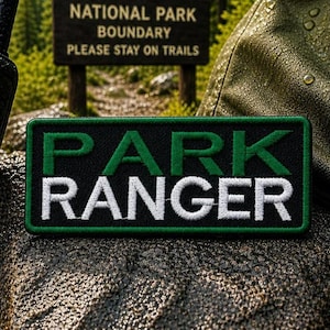 Pode incluir: Um patch retangular com as palavras "PARK RANGER" em letras brancas bordadas sobre um fundo preto, emoldurado por uma borda verde. Um walkie-talkie preto e um uniforme de guarda florestal também são visíveis.