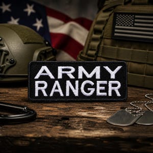 Könnte beinhalten: Ein schwarzer rechteckiger Aufnäher mit den Worten "ARMY RANGER" in weißen Blockbuchstaben. Der Aufnäher wird auf einer Holzoberfläche mit militärischer Ausrüstung und einer amerikanischen Flagge im Hintergrund angezeigt. Erkennungsmarken und ein Karabiner sind ebenfalls sichtbar.