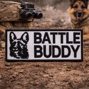 May include: A rectangular patch with a black border and the words "BATTLE BUDDY" in bold, black capital letters. A black and white graphic of a German Shepherd dog is on the left. The patch is on a brown, textured surface.