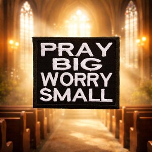 Pode incluir: Um patch bordado preto com as palavras "PRAY BIG WORRY SMALL" em letras brancas. O patch tem uma borda preta. O fundo é um interior de igreja desfocado.