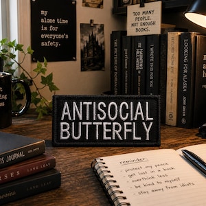 May include: A black patch with white embroidered text that reads "ANTISOCIAL BUTTERFLY" sits on a wooden desk. A black mug with the text "introvert but willing to discuss books" is visible, along with several books and a notebook with handwritten reminders.