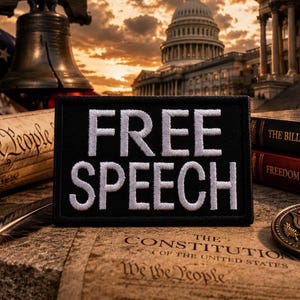 May include: A black patch with the words "FREE SPEECH" embroidered in white. The patch is displayed with the U.S. Constitution, the Bill of Rights, a bronze coin, and the American flag in the background. The U.S. Capitol building is also visible.