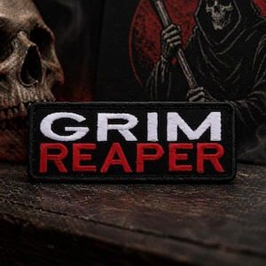 May include: A black rectangular patch with the words "GRIM REAPER" embroidered in white and red. The patch is on a dark wooden surface, with a skull and a Grim Reaper illustration in the background.