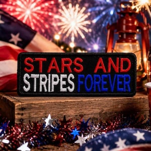 May include: A black patch with the words "STARS AND STRIPES FOREVER" embroidered in red, white, and blue. The patch is displayed on a wooden surface with an American flag and fireworks in the background. Patriotic theme.