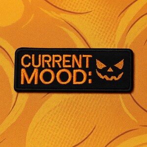 Può includere: Toppa rettangolare nera con le parole "CURRENT MOOD:" in arancione e una faccia di zucca. La toppa è posta su uno sfondo arancione con un motivo ondulato, perfetta per Halloween.