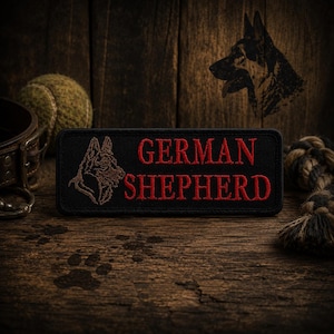 May include: A black rectangular patch with the words "GERMAN SHEPHERD" embroidered in red. A brown German Shepherd dog head is embroidered on the left side. The patch is on a wooden surface with a tennis ball, dog collar, and rope toy.