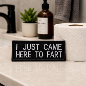 Pode incluir: Um patch retangular preto com o texto branco "I JUST CAME HERE TO FART" é exibido em uma bancada de banheiro. Um rolo de papel higiênico branco e uma pequena planta em vaso também são visíveis.