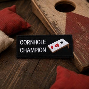 May include: A black patch embroidered with the words "CORNHOLE CHAMPION" and a cornhole board graphic. The patch is surrounded by red and tan bean bags and a cornhole board. The background is a rustic wooden surface.