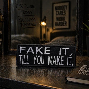 Peut inclure: Un patch noir avec du texte brodé blanc qui dit "FAKE IT, TILL YOU MAKE IT." Le patch est sur une surface sombre, avec un livre et une plante en arrière-plan. Un récipient noir avec le texte "DISCIPLINE FOCUS CONSISTENCY SUCCESS" est à gauche.