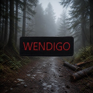 May include: A black rectangular patch with the word "WENDIGO" embroidered in red. The patch is set against a blurred forest backdrop with a path and fog.