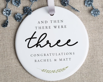 May include: White ceramic ornament with black text that reads "And then there were three. Congratulations Rachel & Matt". The ornament has a green leafy design at the bottom.