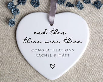 May include: White ceramic heart-shaped ornament with the text "and then there were three" and "Congratulations Rachel & Matt" in black lettering. A small heart is at the bottom of the ornament.