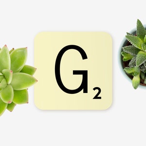 May include: A yellow Scrabble tile with the letter "G" and the number "2" printed on it. The tile is surrounded by two small green succulent plants in pots.