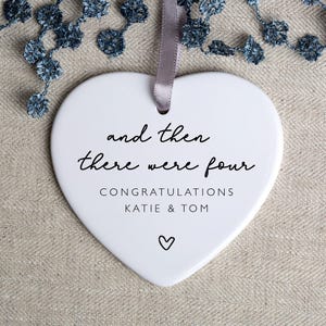 May include: White ceramic heart-shaped ornament with the text "and then there were four" and "Congratulations Katie & Tom" in black lettering. A small heart is at the bottom of the ornament.