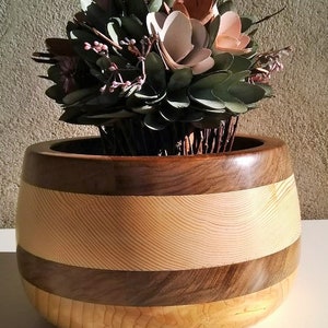 May include: A wooden bowl with a striped pattern in shades of brown and beige. The bowl is filled with a decorative arrangement of faux flowers and greenery.