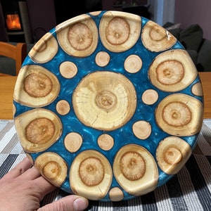 Op de afbeelding: Een ronde houten dienblad met een blauwe epoxyhars achtergrond. Het dienblad is versierd met een cirkelvormig patroon van houten schijven, elk met een uniek houtnerfpatroon.
