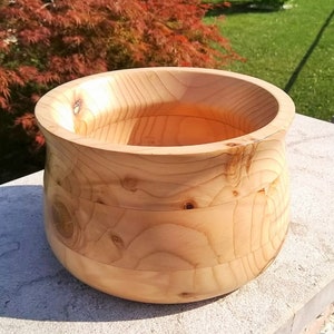 May include: A large, wooden bowl with a natural, light brown finish. The bowl is made of wood with a grain pattern visible on the surface. The bowl is round and has a wide, shallow opening.