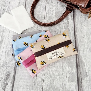 May include: Three rectangular fabric tissue holders featuring a repeating bumblebee pattern in beige, pink, and light blue. Each holder has a horizontal strap and a white label with the black text MISS HEATHCOTE. Two white tissues are visible.