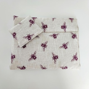 May include: A beige linen fabric pouch with a burgundy bee print. The pouch has a flap closure and a smaller pouch attached to the front.