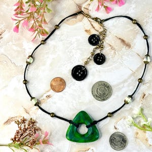 Op de afbeelding: Een handgemaakte ketting met een levendige groene driehoekige hanger met een centraal gat. De ketting heeft een zwart koord, geaccentueerd met kralen en knoopdetails. Decoratieve munten zijn rond de ketting geplaatst.