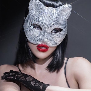 May include: A sparkling silver cat mask with a rhinestone design. The mask covers the eyes and nose, with cutouts for the eyes. The person in the image has red lipstick and black hair. A black lace glove is visible.