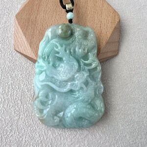 May include: A jade pendant featuring a detailed carving of two dragons intertwined. The pendant is a light green colour and has a smooth, polished finish.