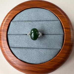 May include: A silver ring with an oval, dark green gemstone. The ring is set in a decorative silver band. The ring is displayed in a wooden jewelry box with a gray interior.