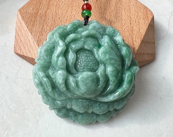 May include: A jade pendant carved in the shape of a flower with a green and white color scheme. The pendant is attached to a brown cord with green and red beads.