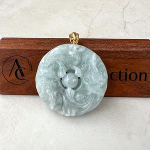May include: A round jade pendant with a gold bail. The pendant is carved with a detailed design of two fish swimming around a central circle. The jade is a light green colour.