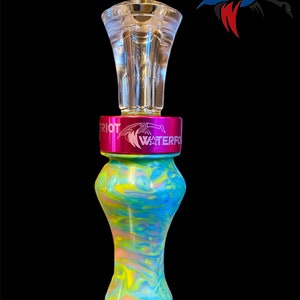 May include: A teal, pink, and yellow marbled duck call with a clear acrylic top and a pink metal band with the words "Patriot Waterfowl" engraved on it.