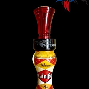 May include: A red and yellow duck call with a black band around the middle. The band has the words "Patriot Waterfowl" engraved on it. The call is decorated with a beer label that says "Premium Grain Belt Beer".