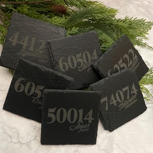 May include: Six black slate coasters with white engraved numbers and city names. The coasters feature the numbers 4412, 60504, 62522, 605, 50014, and 74074. The city names are Lyndhurst, Aurora, Palatine, Osage, Ames, and Stillwater.