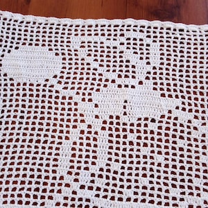 Può includere: Primo piano di una tovaglietta all'uncinetto bianca a trama aperta. La tovaglietta presenta un motivo floreale al centro e un bordo smerlato. Il lavoro all'uncinetto è dettagliato e intricato, con un aspetto delicato e arioso. La tovaglietta è realizzata in morbido filato bianco.
