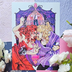 May include: A colourful illustration depicting two women in a dramatic confrontation, one in a red dress, the other in a purple robe. A man in a suit is in the background. The artwork has a theatrical style with bold lines and vibrant colours.