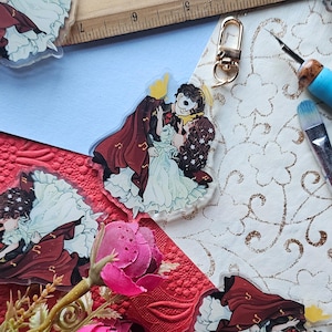 May include: A clear acrylic keychain depicting a dancing couple, inspired by a musical. The man wears a mask and a burgundy cape, while the woman is in a white dress. The keychain has a gold clasp.