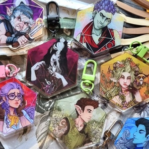 May include: A set of ten colourful enamel pins featuring various characters and designs. The pins are shaped like a traditional twenty-sided die, commonly used in tabletop role-playing games. The pins feature a variety of characters, including a creature with green hair and a crown, a creature with blue hair and a crown, a creature with purple hair and a crown, a creature with red hair and a crown, a creature with yellow hair and a crown, a creature with orange hair and a crown, a creature with pink hair and a crown, a creature with brown hair and a crown, a creature with black hair and a crown, and a creature with white hair and a crown.