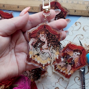 May include: A clear acrylic keychain with a gold clasp, depicting a woman with brown curly hair in a yellow and white dress. The design is framed by a red curtain and roses. Other keychains are also visible.